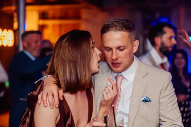 At a lively indoor wedding at Holmes Mill in Clitheroe, a woman with her arm around a man in a beige suit and pink tie talks closely to him, both holding drinks as guests socialize in the background.