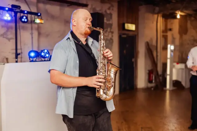 A man plays a saxophone indoors on a wooden floor at Holmes Mill, Clitheroe, eyes closed in concentration. Stage lights and DJ equipment sit behind him, with rustic walls and scattered lighting creating a warm wedding ambiance.