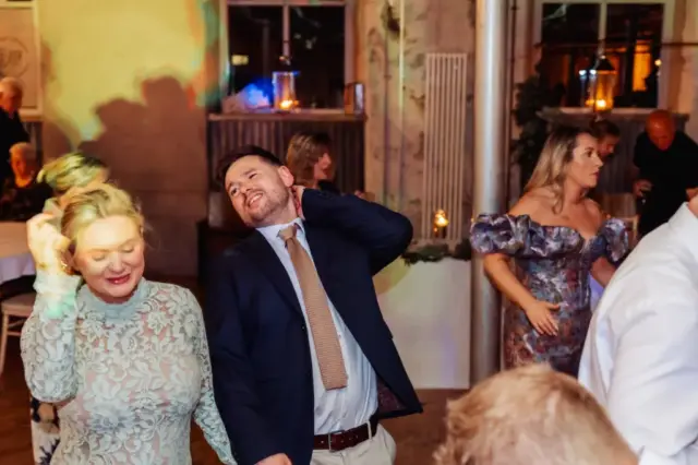 A man in a suit and a woman in a lacy dress dance closely together at a wedding at Holmes Mill, Clitheroe, surrounded by other guests mingling and dancing under warm lighting.
