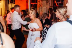 At a Holmes Mill wedding in Clitheroe, guests in semi-formal attire dance and enjoy themselves. A woman in a white dress holds a drink and sings along, surrounded by lively partygoers at this joyful indoor celebration.