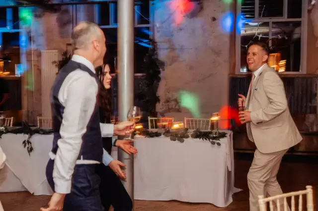 Three people dressed in formal attire are dancing at a Holmes Mill wedding in Clitheroe, with colorful lights and a decorated table with candles in the background. One person holds a drink and smiles enthusiastically.