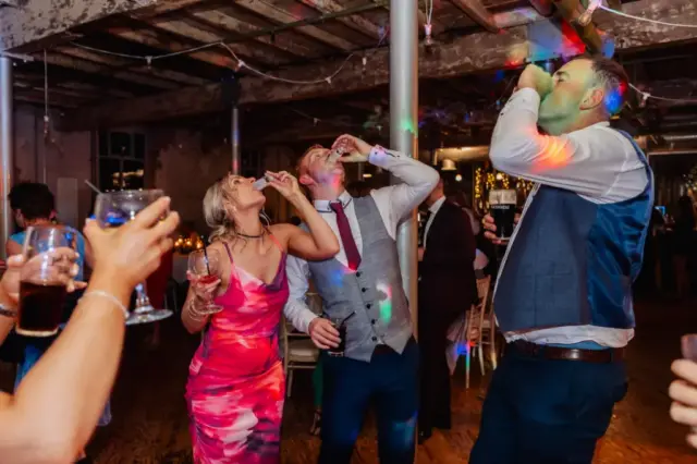 At a lively Holmes Mill wedding in Clitheroe, three people—one woman in a pink dress and two men in suits—celebrate on the dance floor taking shots, surrounded by colorful lights and joyful guests.