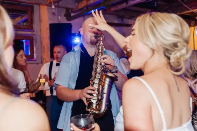 A lively wedding party scene at Holmes Mill, with a saxophonist playing as a woman in a white dress joyfully raises her hand near his face. Other guests are visible in the background, enjoying the festive Clitheroe atmosphere.
