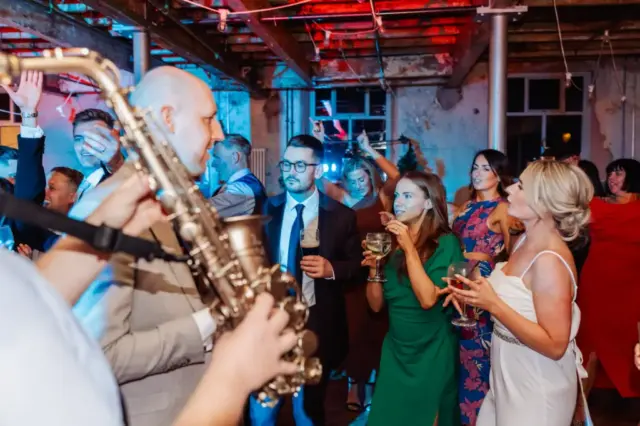 Guests in formal attire dance and enjoy drinks at an indoor wedding party at Holmes Mill, Clitheroe, as a saxophonist plays in the foreground under vibrant, colorful lights.