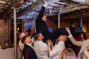 At a lively Holmes Mill wedding in Clitheroe, a group of men in suits lift another man above their heads as other guests watch and smile in the background.