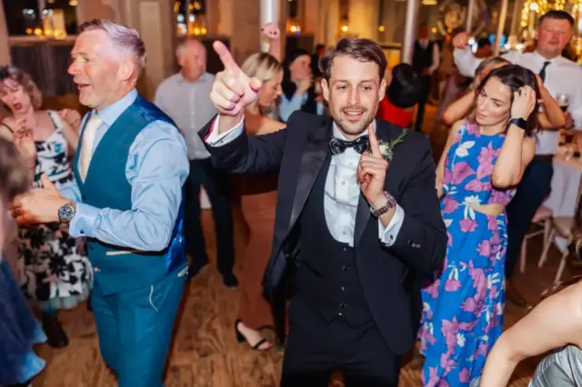 A group of people in formal attire dance and celebrate at a Wedding reception at Holmes Mill, Clitheroe, with a man in a tuxedo pointing upward in the foreground.