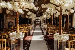 An elegant indoor wedding aisle lined with gold chairs and tall artificial white blossom trees, leading to a decorated altar accented by carton towers under warm, ornate lighting in a grand, richly decorated hall.