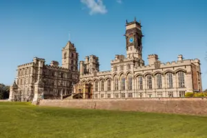 A large historic stone building with arched windows, towers, and a clock tower—Carton Towers—set against a clear blue sky and surrounded by a grassy lawn; an ideal backdrop for a romantic wedding.