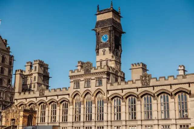 A historic stone building with tall arched windows and intricate detailing, featuring a central clock tower reminiscent of carton towers, with a blue clock face, set against a clear blue sky—an enchanting spot for a wedding.