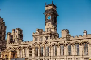 A historic stone building with tall arched windows and intricate detailing, featuring a central clock tower reminiscent of carton towers, with a blue clock face, set against a clear blue sky—an enchanting spot for a wedding.