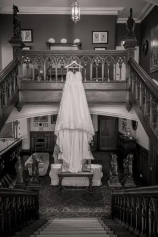 A wedding dress hangs from a balcony railing in a grand, vintage interior with ornate wooden staircases and classic decor, captured in black and white at Carton Towers.