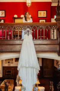 A smiling bride in a white robe stands on a wooden balcony, looking down at her wedding dress, which hangs from the railing in a richly decorated room with red walls, framed art, and views of distant carton towers—ready for her dream wedding.