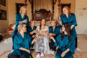 A bride in a white robe sits on a couch, surrounded by six bridesmaids in matching teal robes. They are indoors at a wedding, smiling and toasting with champagne glasses in a warmly decorated room beside carton towers.