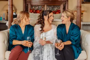 Three women sit together on a sofa, smiling and holding champagne glasses at a wedding. The woman in the middle wears a white lace dress, while the two on either side wear teal robes. The atmosphere is joyful and celebratory.
