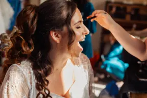 A woman with long, curly brown hair, wearing a white lace robe, smiles with her mouth open as another person applies wedding makeup to her face in a brightly lit room.
