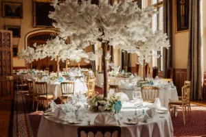 A lavish banquet hall set for a wedding, featuring round tables with tall white artificial floral centerpieces resembling carton towers, surrounded by gold chairs and bathed in natural light from large windows.