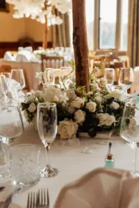 A close-up of an elegant wedding table setting with clear glassware, plates, and silverware, featuring a centerpiece of white roses and greenery on a white tablecloth in a softly lit room.