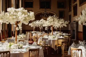 Elegant banquet hall set for a wedding, with round tables featuring gold chairs and tall white flower centerpieces, under soft natural light from large windows. Paintings, a chandelier, and striking carton towers complete the sophisticated setting.