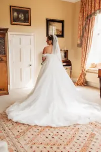 A bride in a white wedding gown and veil stands facing a closed white door in an elegant room with patterned curtains, carton towers, framed pictures on the wall, and sunlight streaming in through a window.
