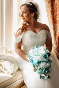A bride in a white, off-shoulder wedding dress with beading sits by a window at Carton Towers, holding a bouquet of white and blue flowers. She gazes outside, sunlight illuminating her face and hair on her wedding day.