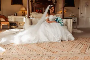 A bride in a white, off-the-shoulder wedding dress and veil sits on a sofa in an elegant, warmly lit room with carton towers in the background, holding a bouquet of blue and white flowers, smiling and looking to the side.
