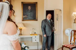 A man in a suit smiles warmly as he enters a room where a bride in a white dress stands, holding a bouquet. The elegant decor includes a desk, bottles of water stacked like carton towers, and a portrait painting, setting the perfect wedding scene.