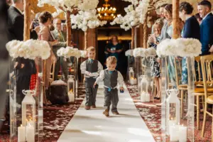 Two young boys in matching vests and bow ties walk down a flower-petal-covered wedding aisle, flanked by white floral arrangements, lanterns, and decorative carton towers, while guests look on from both sides.