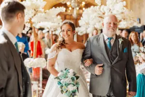 A smiling bride in a white dress with a bouquet walks down the aisle at her wedding, accompanied by an older man in a gray suit; carton towers and white floral decorations add charm as guests look on and a man in a suit waits at the front.