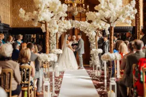 A bride and groom stand under a floral arch, holding hands during their wedding ceremony in an ornate hall, surrounded by guests and elegant white flower arrangements lining the aisle beneath grand carton towers.