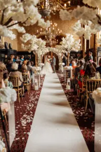 A bride and groom stand at the altar under white floral arches in an ornate, gold-accented wedding room, surrounded by guests along a white aisle sprinkled with flower petals, elegant lanterns, and decorative carton towers.