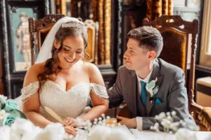 A bride in a white dress smiles while signing a wedding document, sitting next to a groom in a gray suit who looks at her affectionately. They are indoors, seated at a decorated table with flowers and carton towers as unique decor.