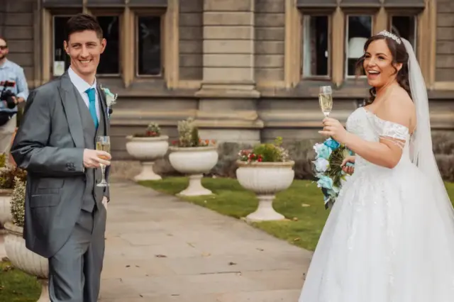 A smiling bride in a white gown and a groom in a gray suit celebrate their wedding outdoors with glasses of champagne, surrounded by potted plants, green grass, and the charming backdrop of stone buildings and carton towers.