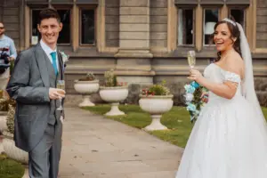 A smiling bride in a white gown and a groom in a gray suit celebrate their wedding outdoors with glasses of champagne, surrounded by potted plants, green grass, and the charming backdrop of stone buildings and carton towers.