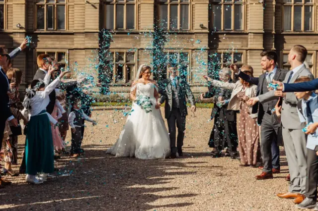 A bride and groom walk outside a grand building with carton towers as guests in formal attire shower them with blue confetti, celebrating their wedding.