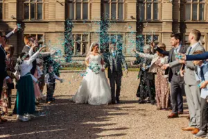 A bride and groom walk outside a grand building with carton towers as guests in formal attire shower them with blue confetti, celebrating their wedding.