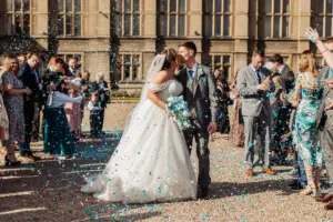 A bride and groom share a kiss outdoors at their wedding as guests throw blue confetti. The bride wears a white dress and veil, the groom a grey suit. Carton Towers stands grandly in the background.