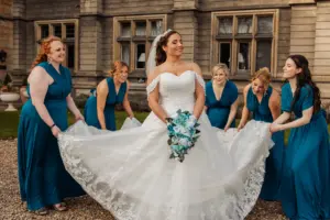 A bride in a white gown holds a bouquet and smiles, surrounded by five bridesmaids in teal dresses who are lifting the train of her dress outside the grand stone carton towers on her wedding day.