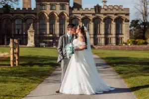 A bride and groom share a wedding kiss on a pathway outside the grand Carton Towers. The bride wears a white gown and holds a bouquet; the groom is in a gray suit. Sunlight, green lawns, and blue sky complete the romantic scene.