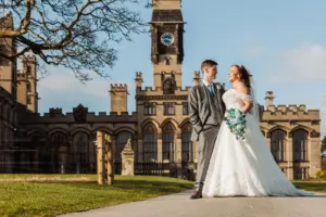 A bride and groom stand together, smiling at each other, in front of a large historic wedding venue with carton towers and a grand clock tower. The bride holds a bouquet of blue and white flowers as the sun casts warm light on their special day.