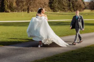 A bride in a white gown lifts her dress as she walks on a path at an outdoor wedding, smiling at a man in a gray suit and blue tie. They are surrounded by green grass, trees, and distant carton towers under the sunny sky.