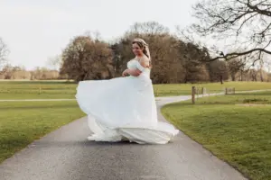A bride in a white wedding dress twirls on a paved path outdoors, surrounded by green grass, carton towers, and leafless trees under a bright sky.