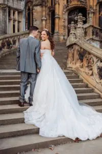A bride in a white gown and a groom in a gray suit stand hand in hand on stone steps, smiling back at the camera, with an ornate historic building and distinctive carton towers in the background, capturing their wedding day.