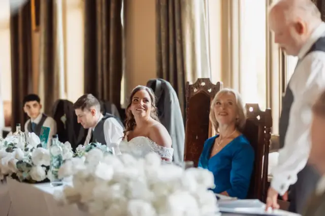 A bride in a white dress smiles at an older man giving a speech at a wedding table, while a woman in blue and other guests sit beside her. The elegant room features large windows, floral arrangements, and carton towers as unique décor accents.