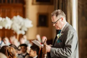 A man in a suit with a boutonnière reads from papers at a formal wedding indoors, with guests seated and blurred in the background, perhaps near the elegant carton towers.