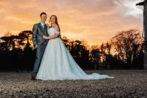 A bride in a white wedding dress and a groom in a gray suit stand together outdoors, smiling at their wedding, with carton towers and trees silhouetted against a dramatic sunset sky in the background.