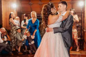 A bride and groom share their first dance at the wedding, surrounded by guests. The bride’s white gown and groom’s gray suit stand out as children and women in colorful dresses watch among carton towers in a warmly lit room.