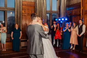A bride and groom share a first dance in an elegant room with wooden walls and large windows at their wedding, surrounded by guests watching and taking photos near the beautiful carton towers.