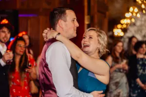 A smiling couple dances closely at a wedding celebration, surrounded by guests in festive attire. Warm lighting and blurred background figures—plus towering cartons—create a joyful, lively atmosphere.
