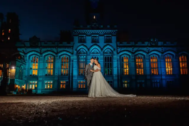 A bride and groom stand together in front of the grand, illuminated Carton Towers at night, the windows glowing warmly as blue lights highlight the building’s detailed architecture behind them.