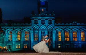 A bride and groom share a romantic wedding dance at night in front of the illuminated arched windows of historic Carton Towers, beautifully bathed in blue lighting.
